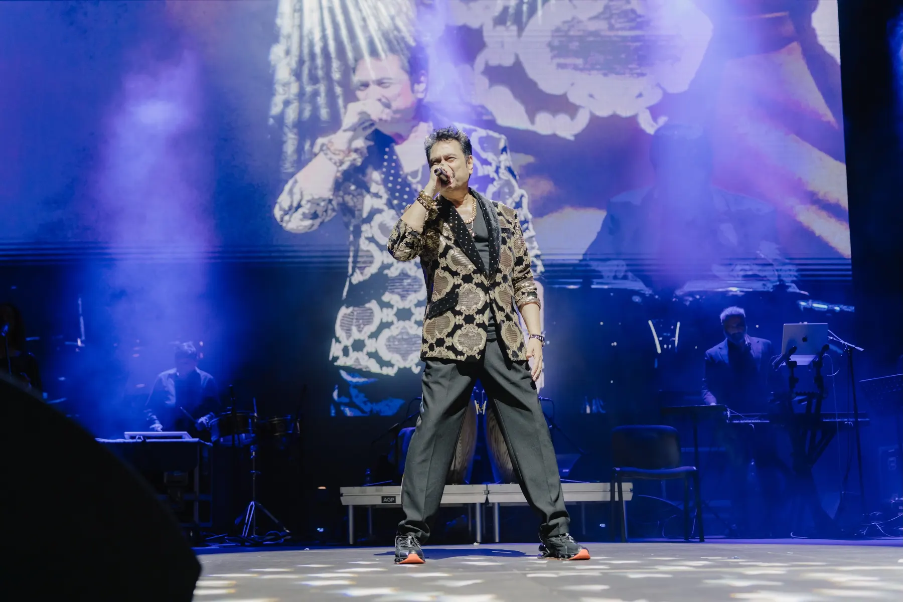 Kumar Sanu singing live on a concert stage in front of a giant screen and his band